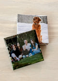 A small stack of photo magnets displayed on a wooden surface, one with a family and the other with a dog in the snow.