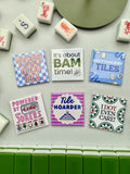 A set of square magnets featuring humorous mahjong-themed quotes and designs, arranged on a light gray surface with a row of green and white mahjong tiles at the bottom.