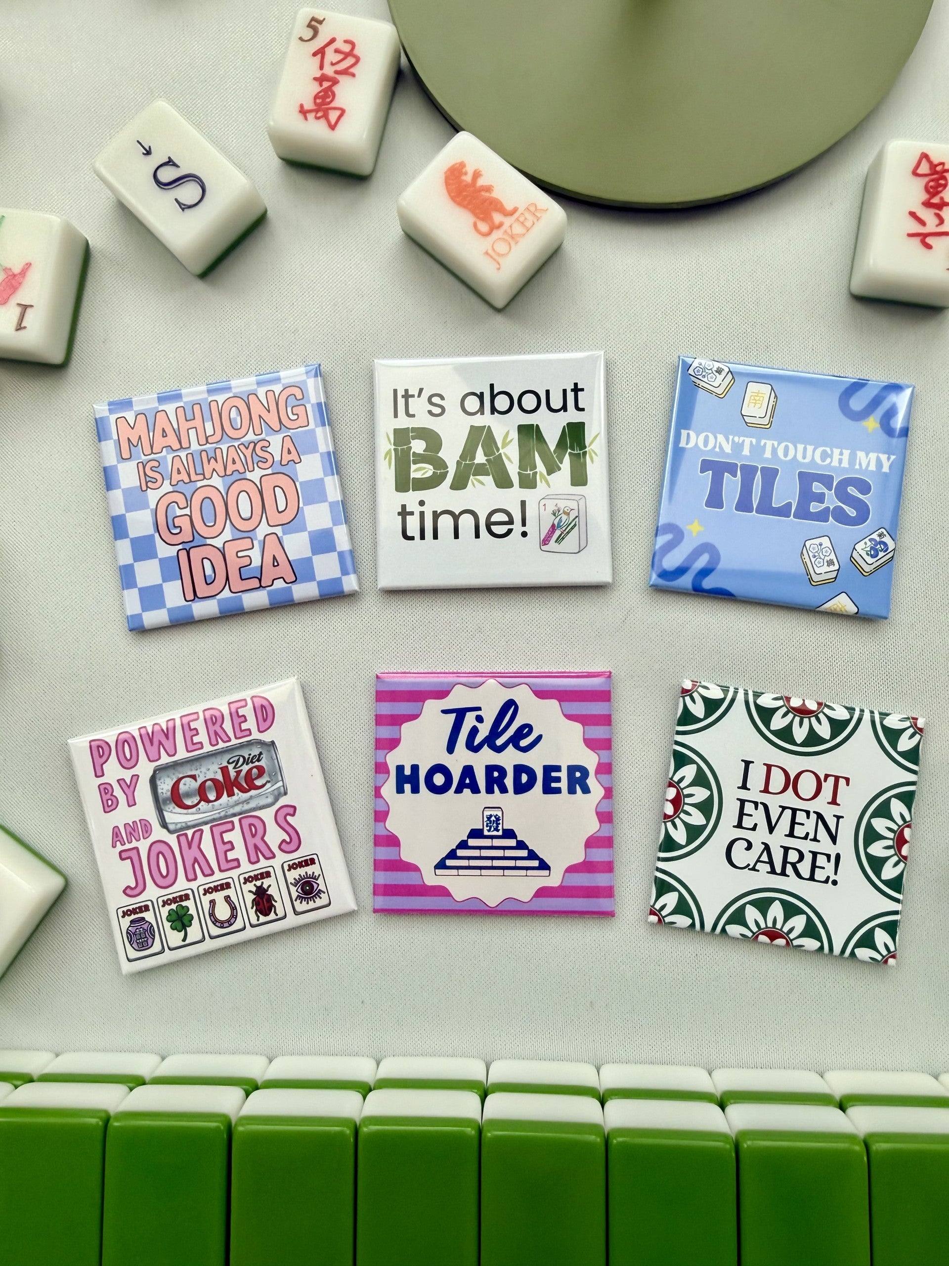 A set of square magnets featuring humorous mahjong-themed quotes and designs, arranged on a light gray surface with a row of green and white mahjong tiles at the bottom.