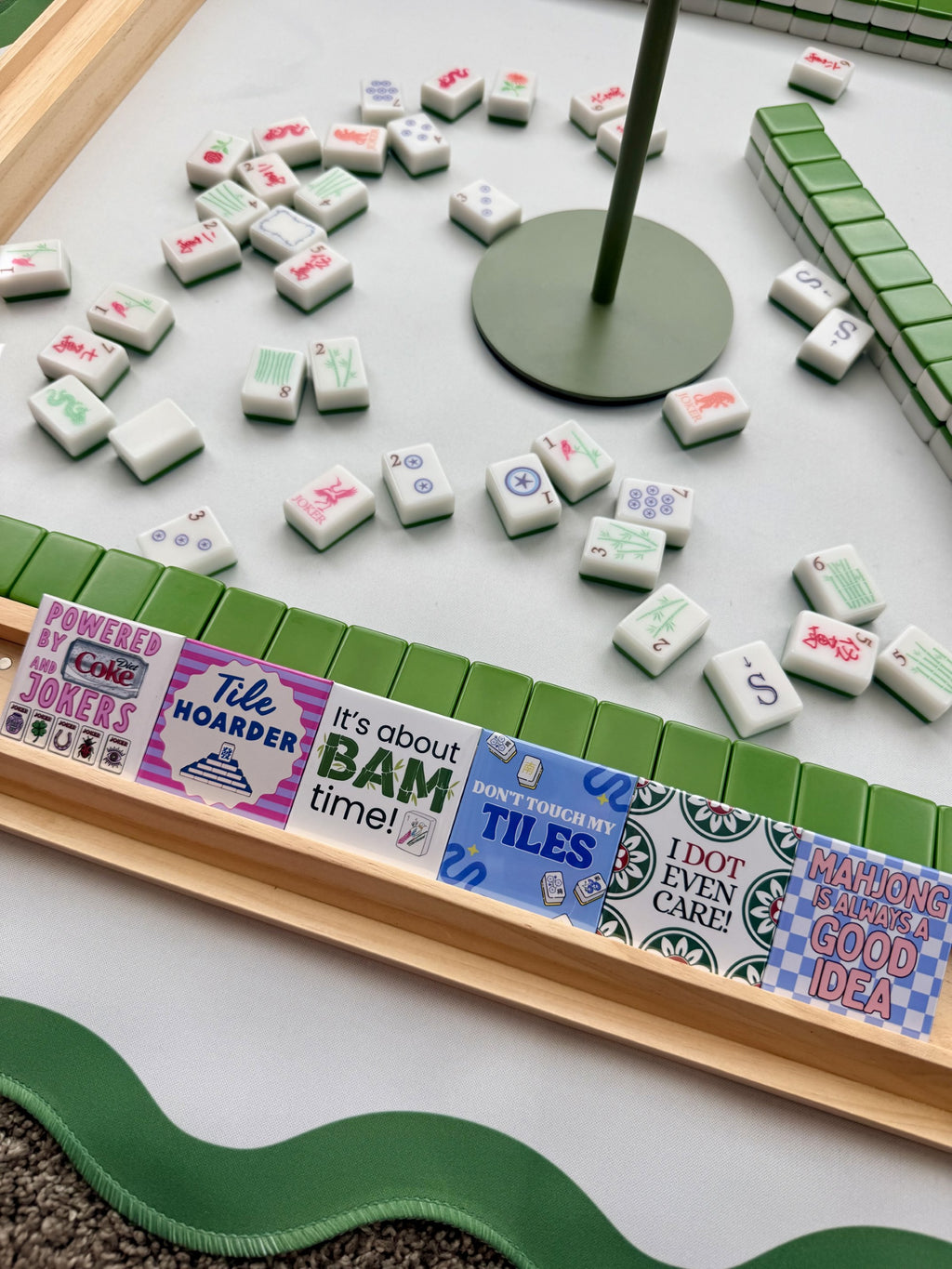 A close-up of a Mahjong game board with white tiles scattered across a light gray surface, green tile racks, a wooden frame, and decorative printed tiles with phrases like 'Tile Hoarder' and 'BAM time!'.